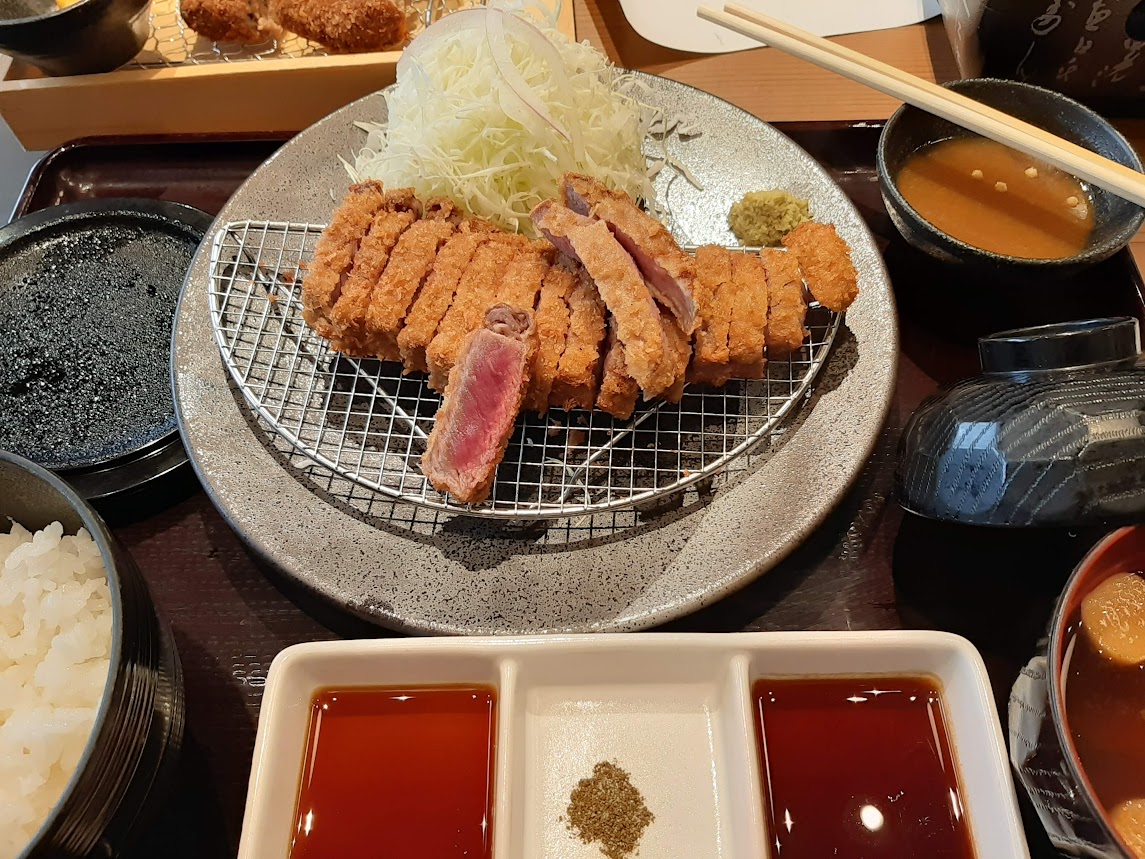 中央に山盛りキャベツの千切りと牛カツ。一切れだけレアな断面を見せている。味噌汁とごはん。手前にタレらしきものとワサビの四角い皿。全国展開する牛カツ専門店「牛カツ京都勝牛」の牛カツ膳