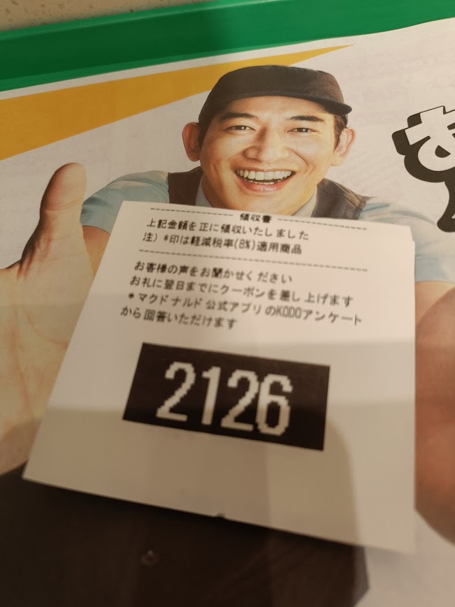 マクドナルドのクルーが笑顔で手を差し伸べるポスターの前に、裏返されたレシートが置かれている。レシートには黒地に白抜きの大きな数字「2126」が印字されている。