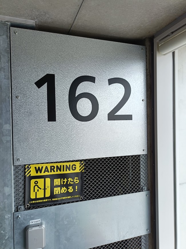 メタリックな壁面に設置された、大きな黒い数字「162」のプレート。その下には黄色の「WARNING 開けたら閉める!」というステッカーとインターホンがある。